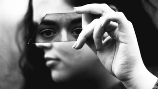 Black and white profile of woman covering her eyes with a mirror that shows front perspective of eyes